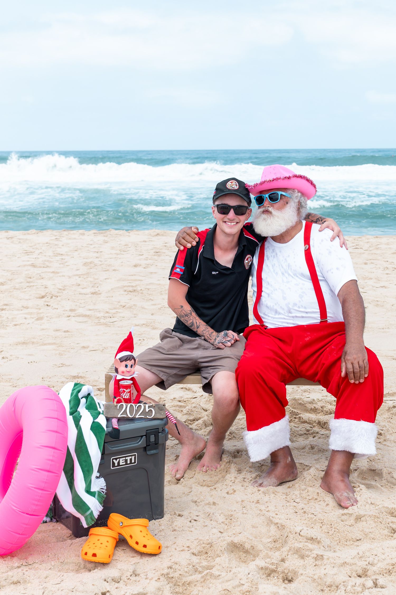 Two people relax on a sandy beach with colorful inflatables and a Yeti cooler near the ocean waves.
