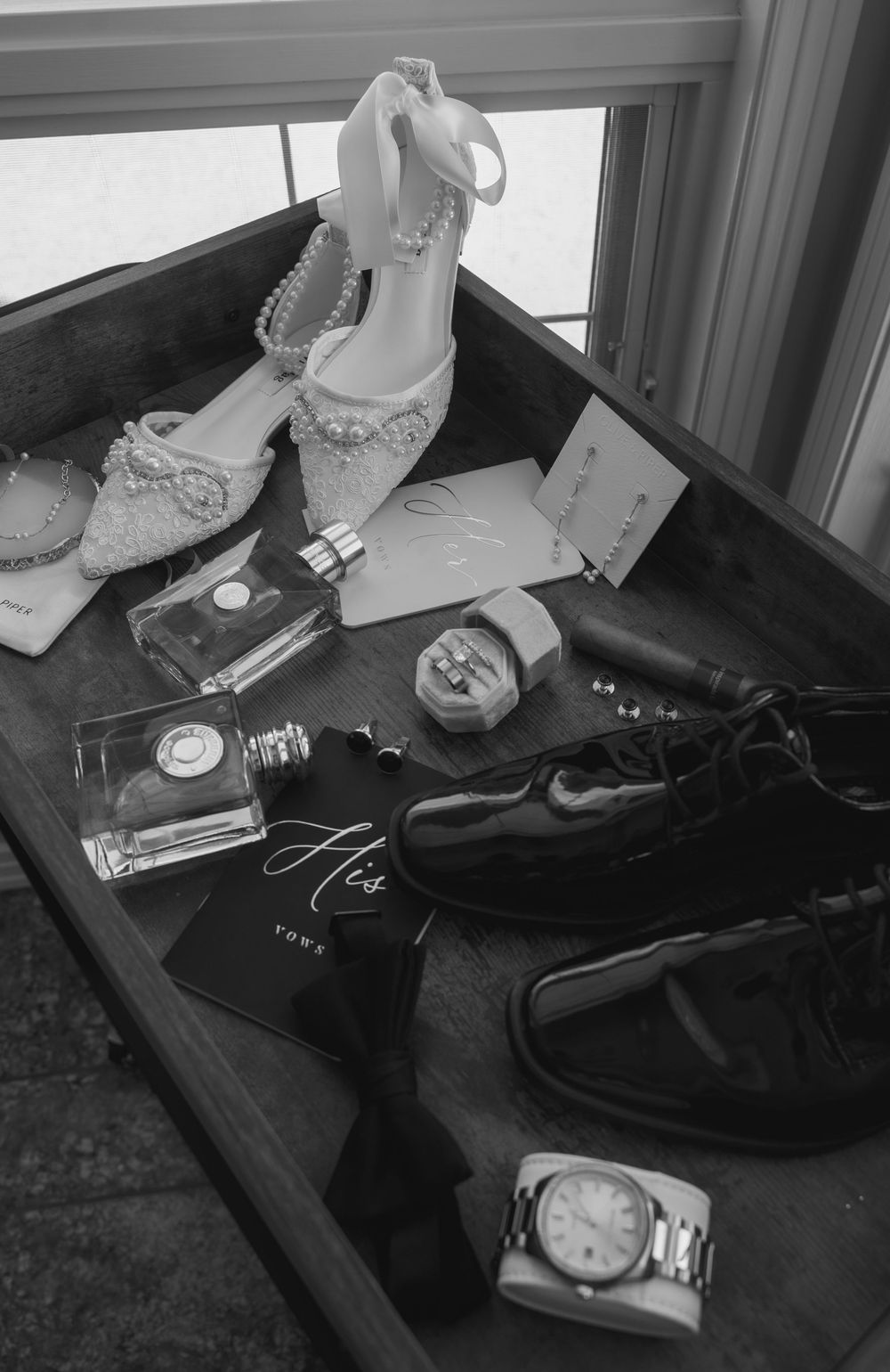 Black and white photo of wedding day accessories and details arranged on wooden surface.