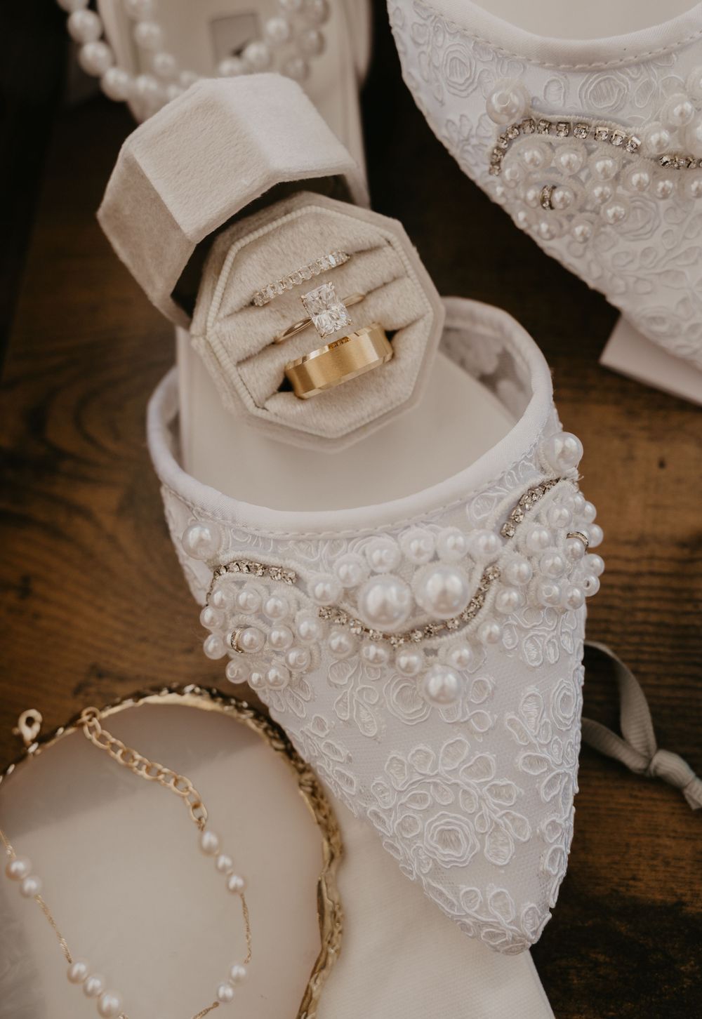 Delicate white bridal shoes adorned with pearls and lace details on wooden surface.