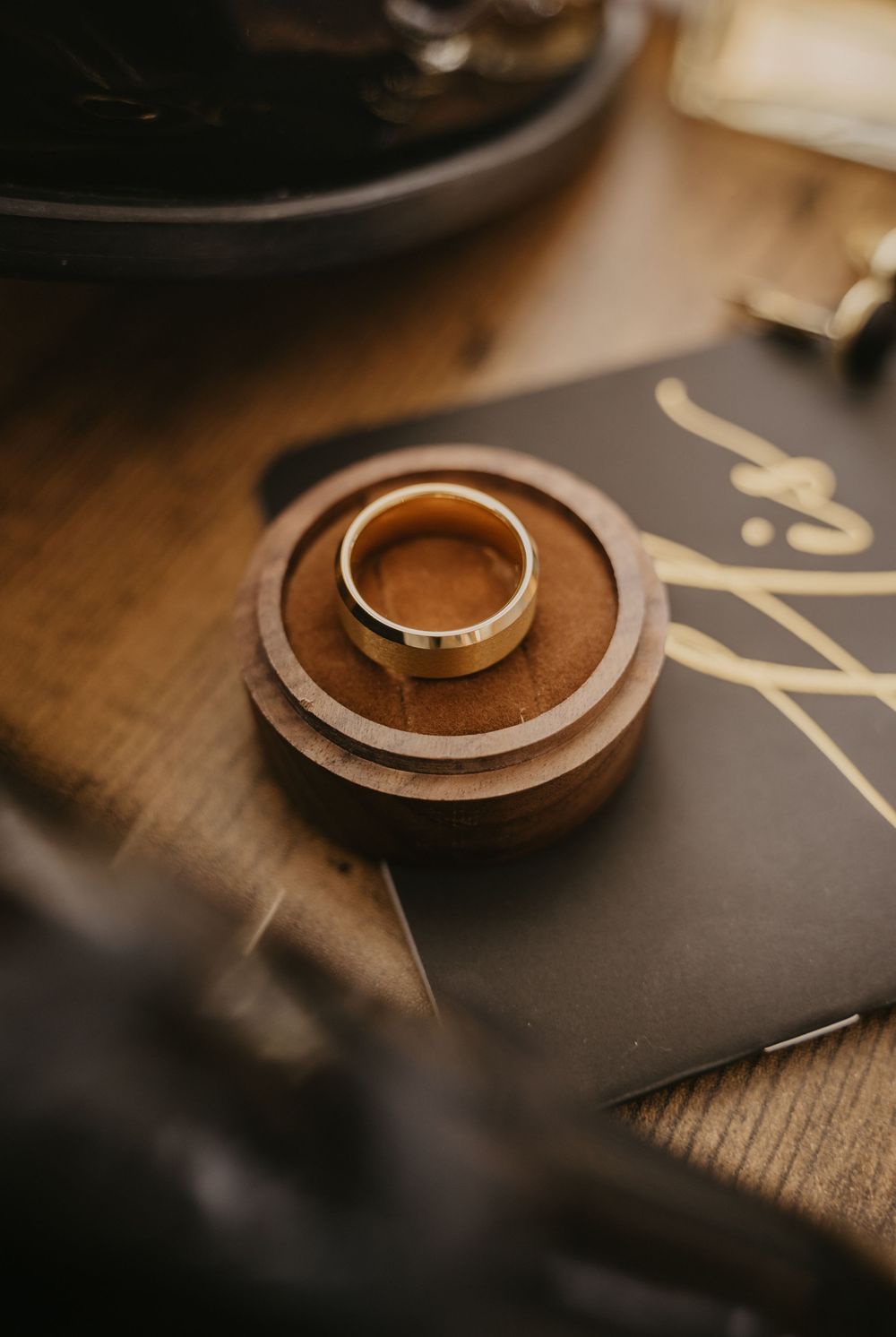 A silver wedding ring sits on a small wooden dish atop a dark surface in warm, moody lighting.