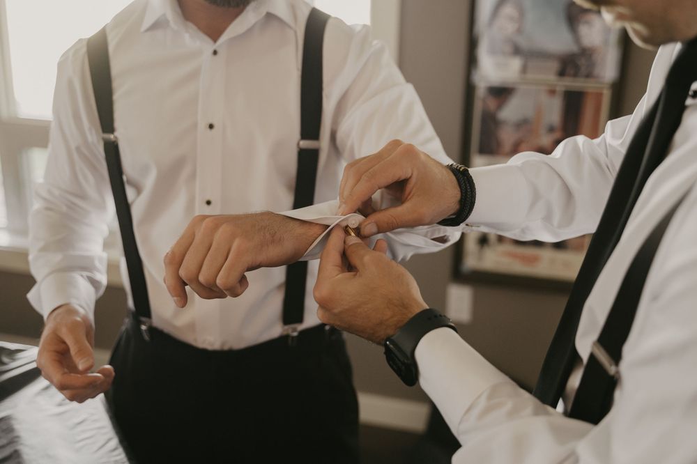 Close up of groomsmen in white shirts and suspenders helping each other get ready.