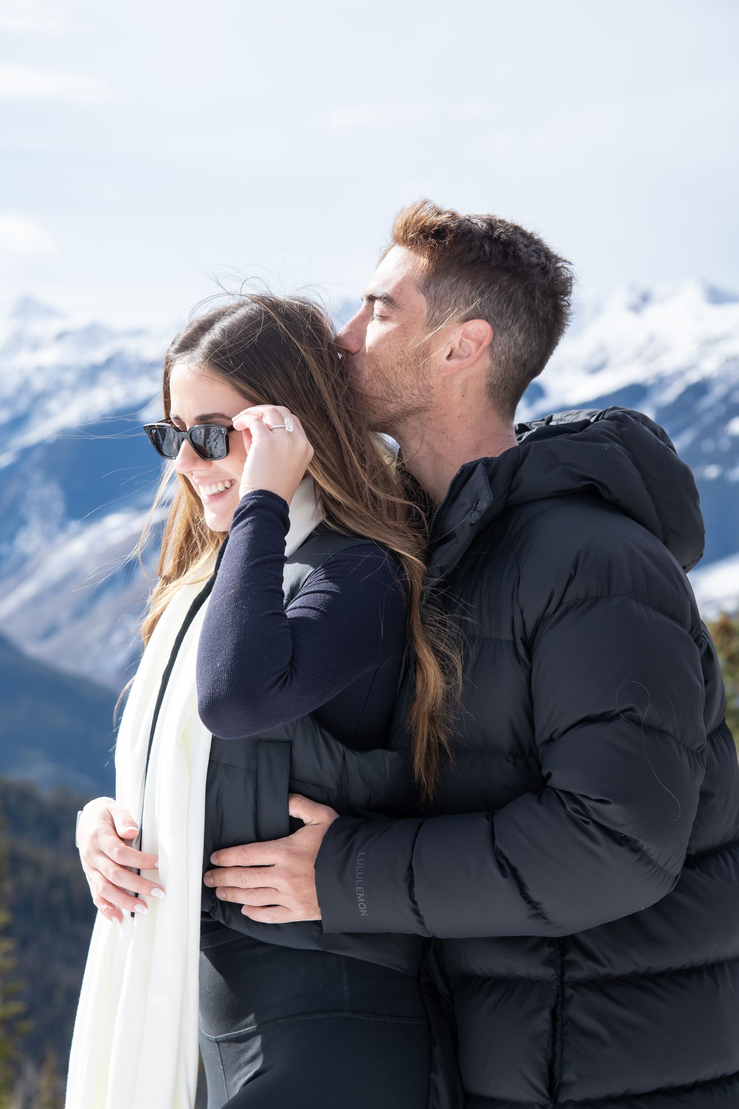 Stylish couple stand closely on top of Aspen mountain, the girl holds her sunglasses and looks down after a surprise proposal