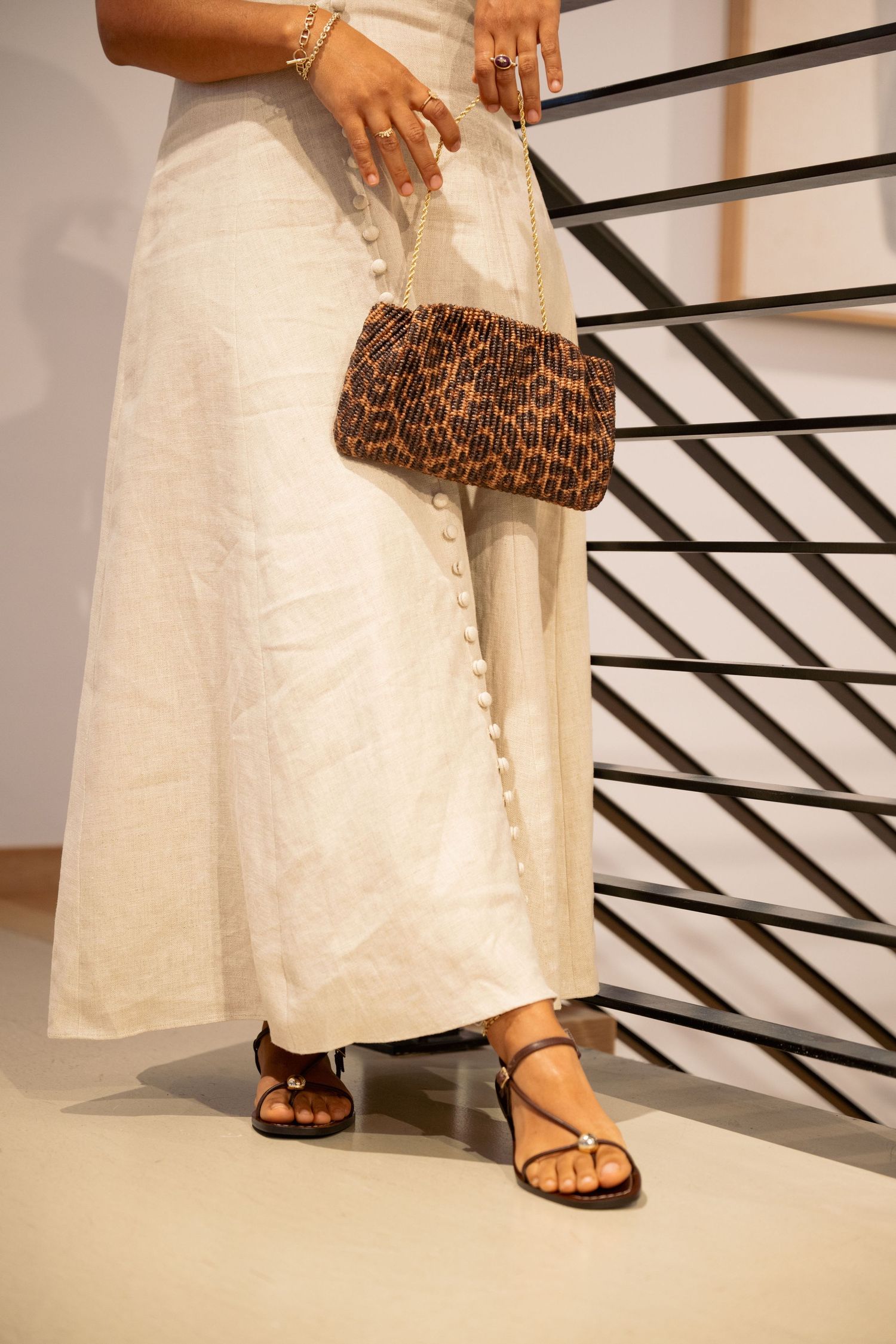 A person wearing a flowing white dress and brown sandals stands against a modern interior with black railings.