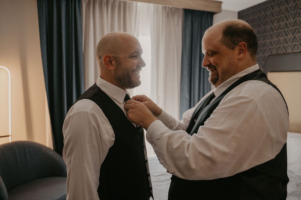 Wedding preparation scene showing formal attire adjustments in natural window light.