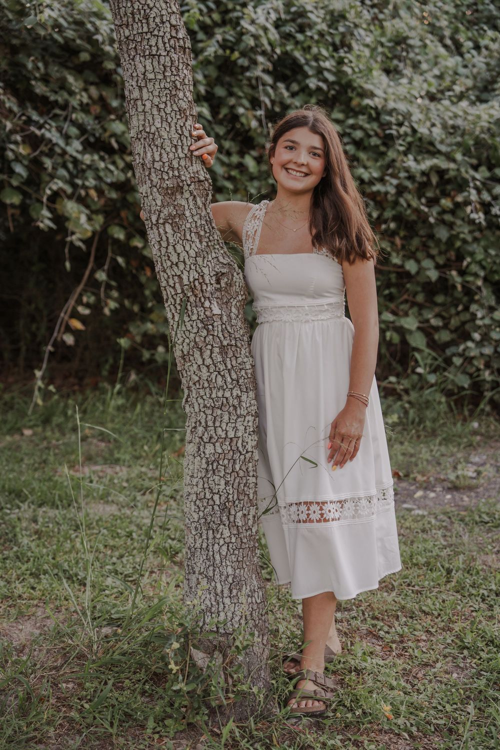 Young person in white summer dress posing by tree trunk in outdoor setting.