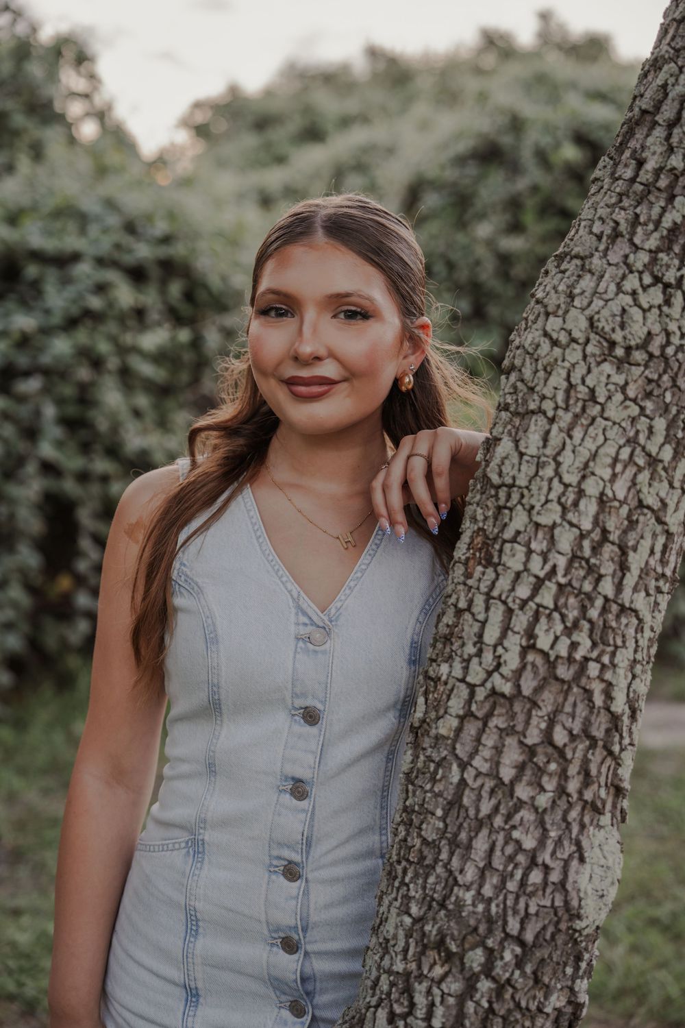 Portrait of someone in a light denim button-up dress leaning against a tree trunk in a natural outdoor setting.