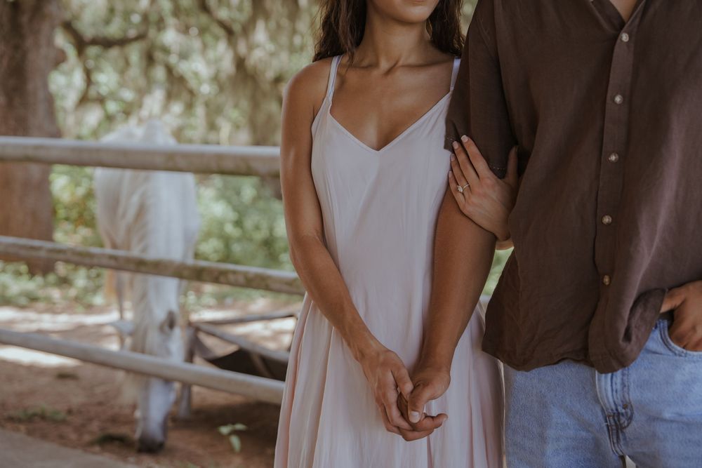 Couple standing together in casual attire near a wooden fence on a sunny day.