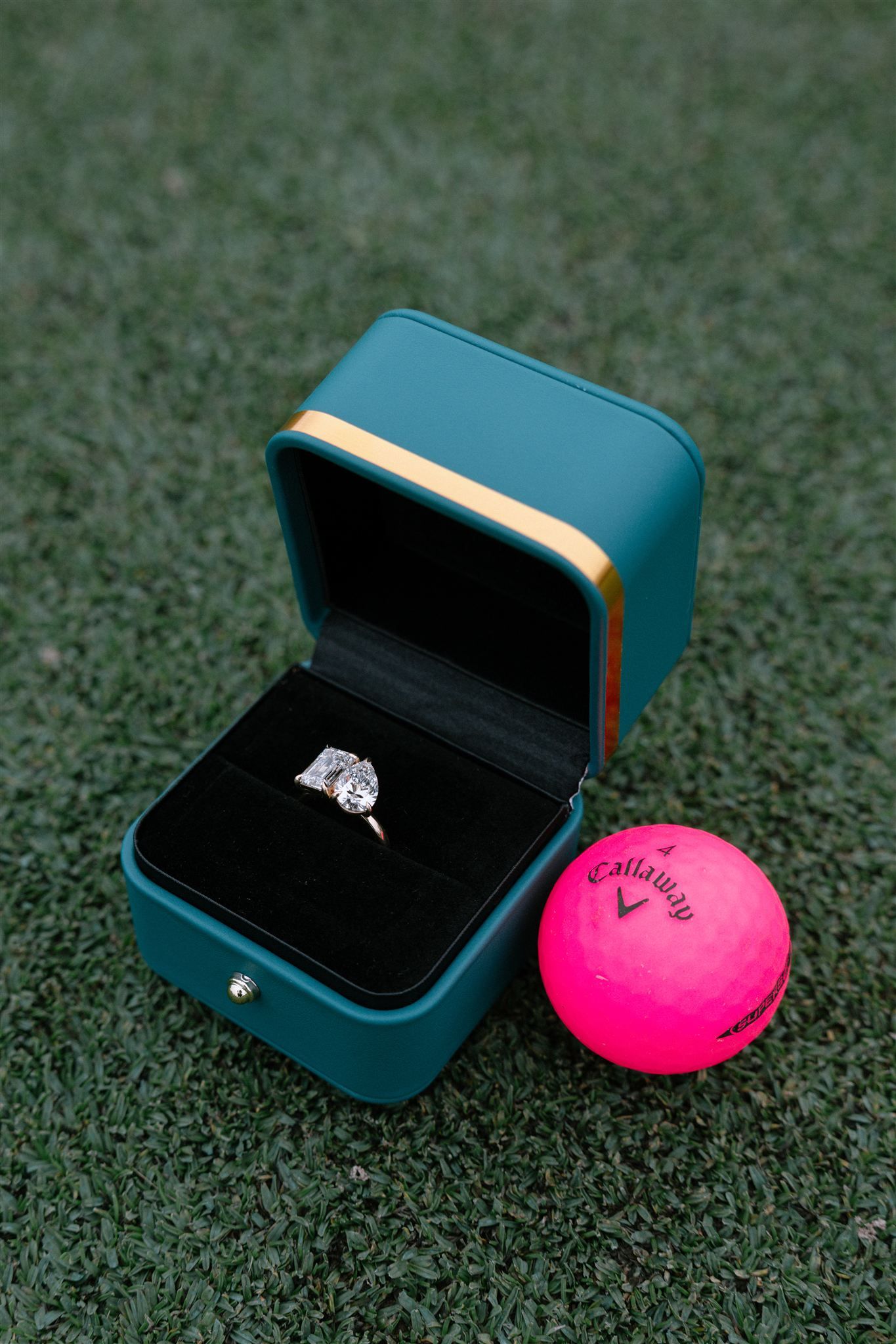 Diamond engagement ring in teal velvet box next to bright pink Callaway golf ball on green turf surface.