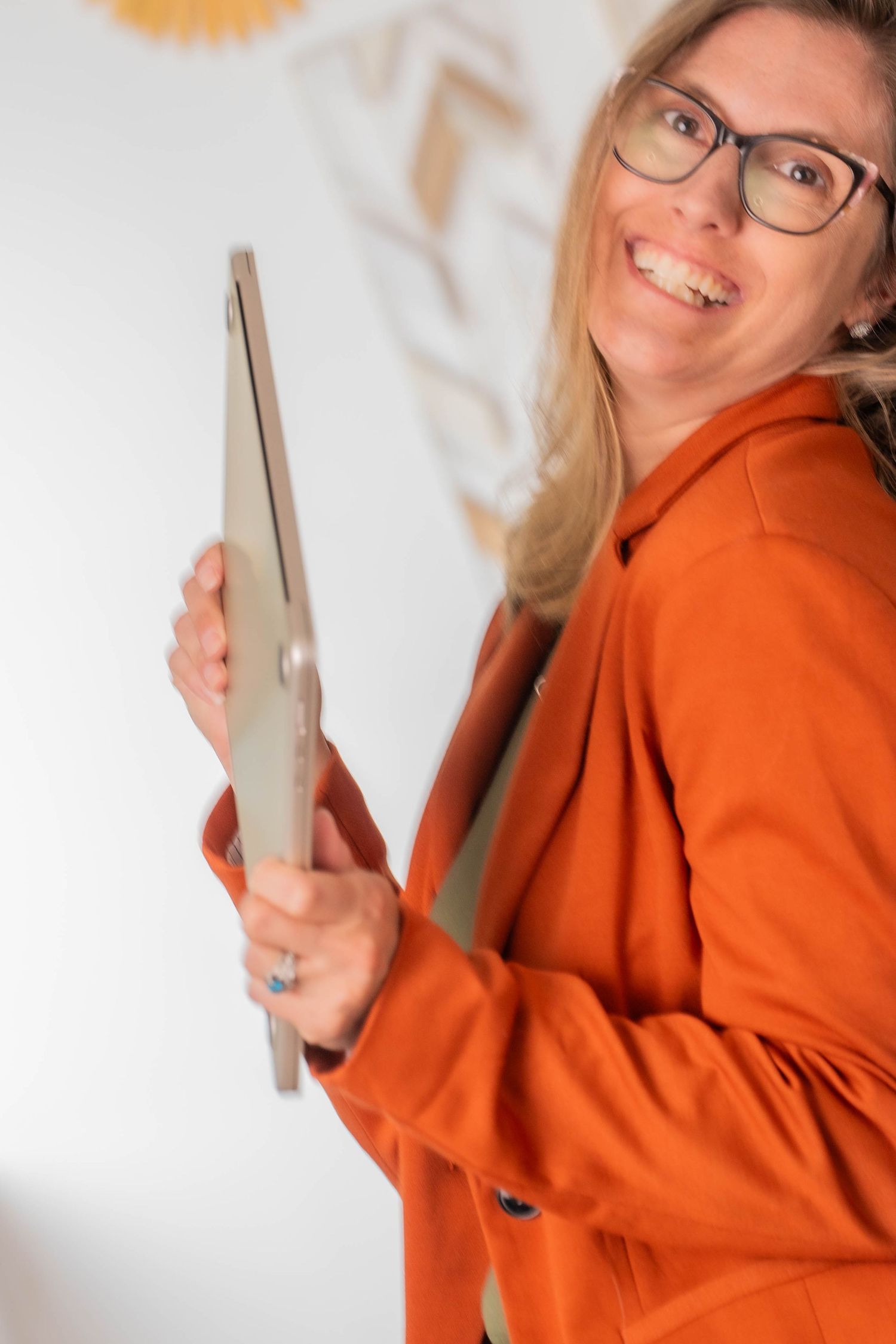 NH photographer in orange blazer holding laptop with playful expression against white wall.