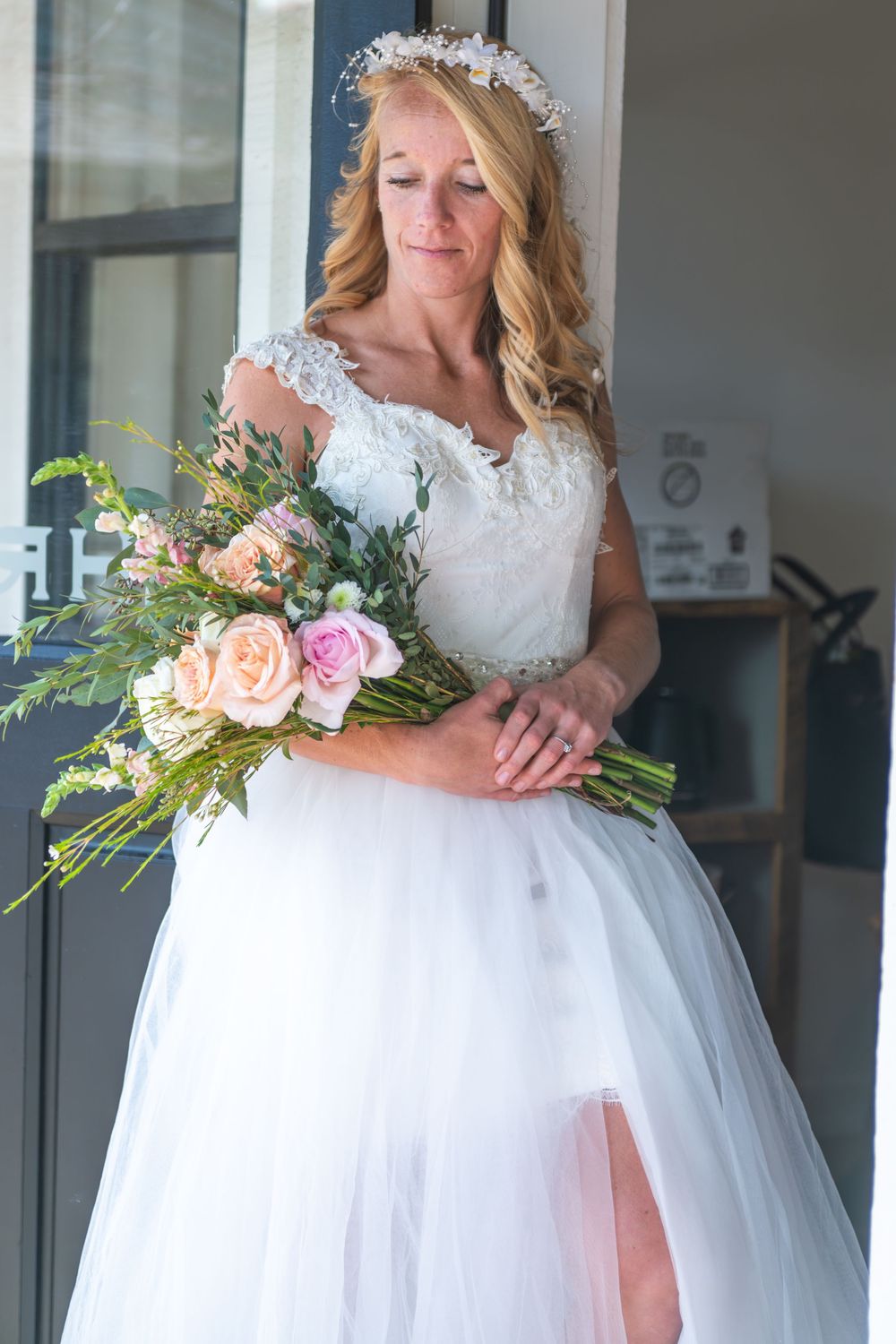 A bride in a white lace gown with tulle skirt holds a romantic bouquet of pink roses and greenery in a doorway in natural light.