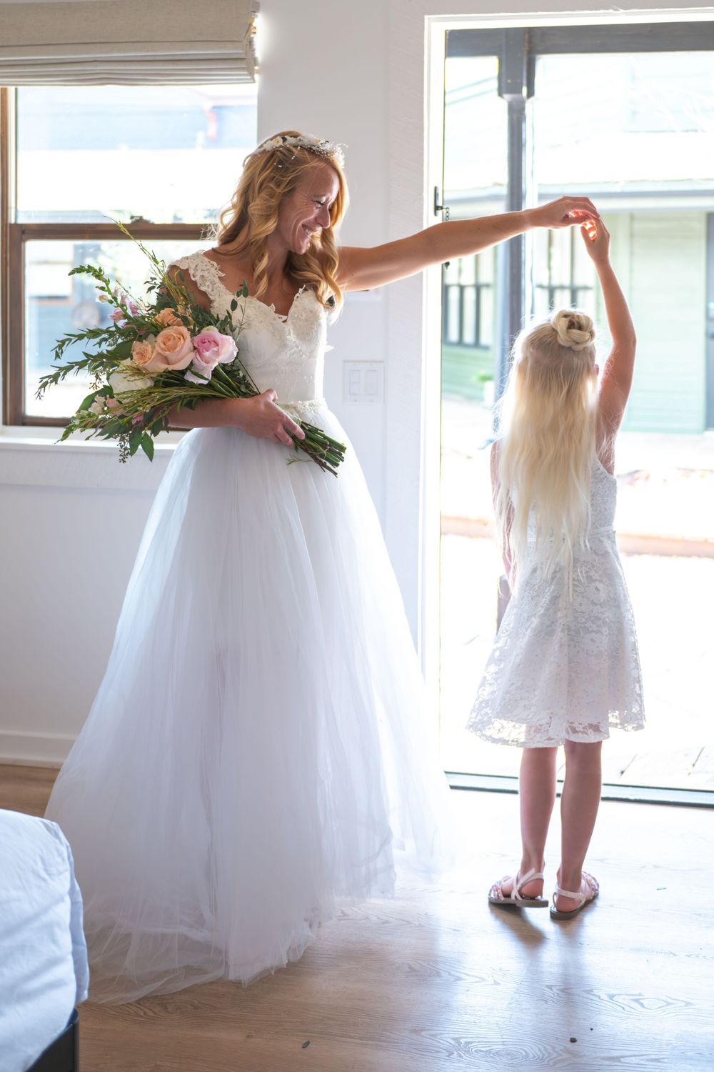 A bride is dancing with her daughter in a natural light. The bride is holding a bouquet, the daughter is holding her mother's hand and spins