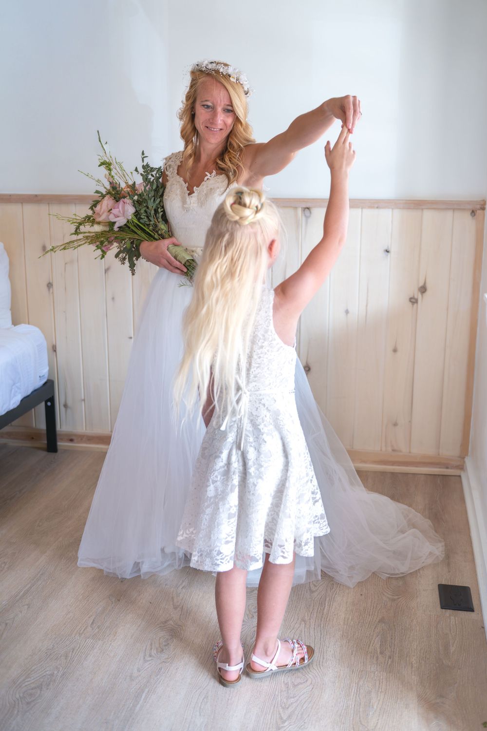 A bride is dancing with her daughter in a natural light. The bride is holding a bouquet, the daughter is holding her mother's hand and spins