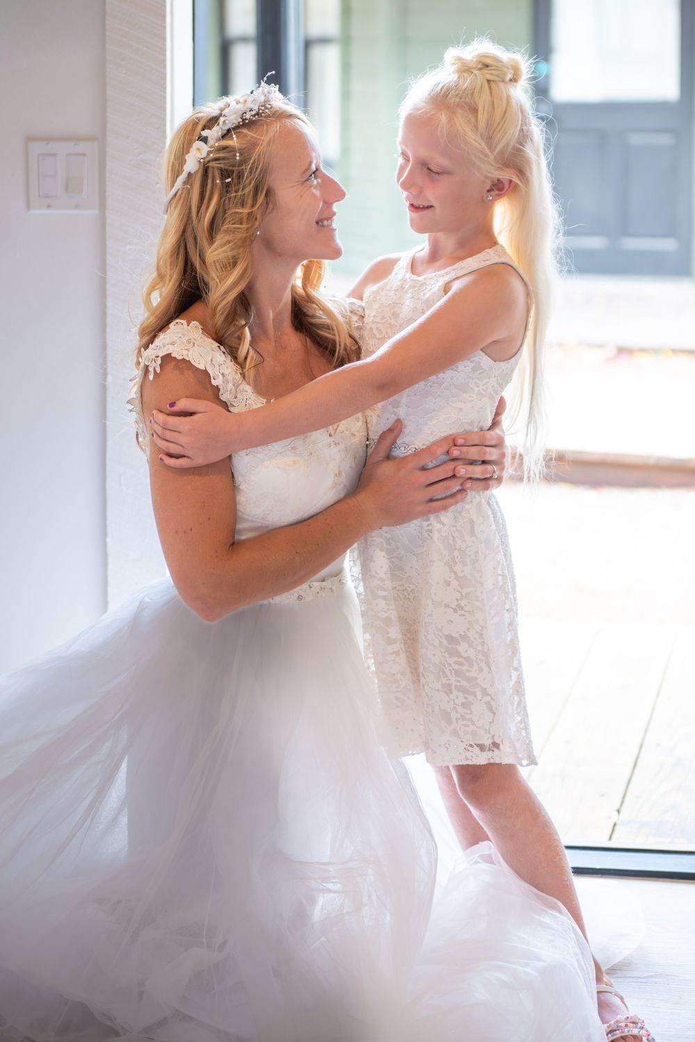 A bride is kneeling hugging her daughter. Both are in white lace dresses in a door with natural light coming in. They smile at each other.