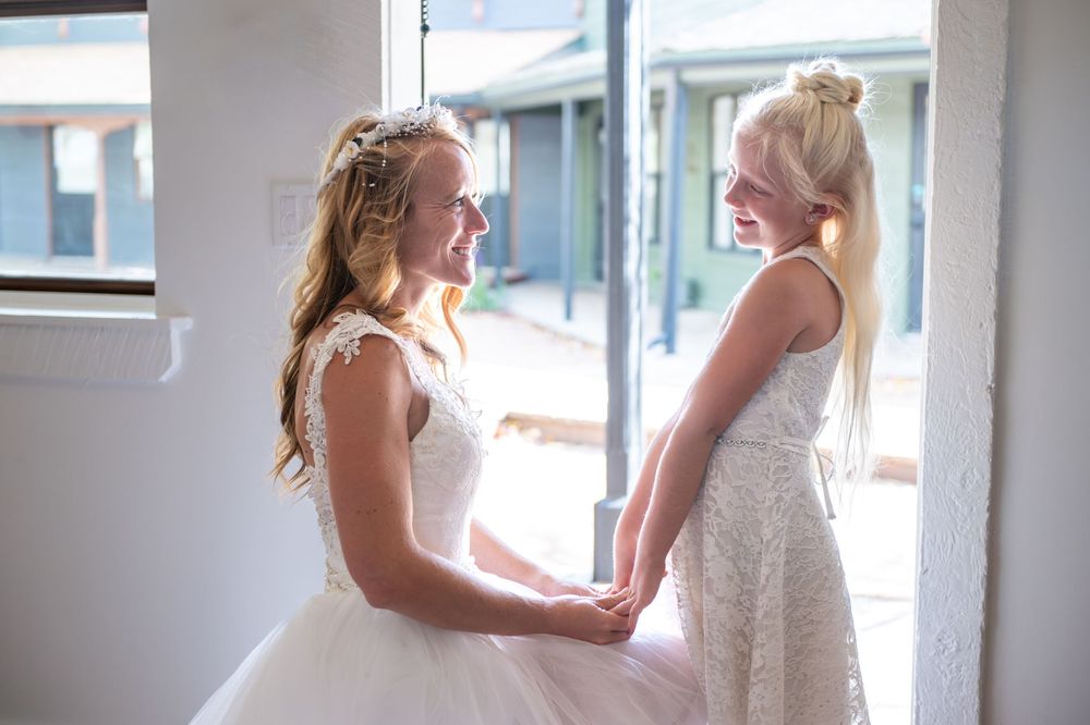 A bride is kneeling holding both of her daughter's hand. Both are in white lace dresses in a door with natural light coming in.