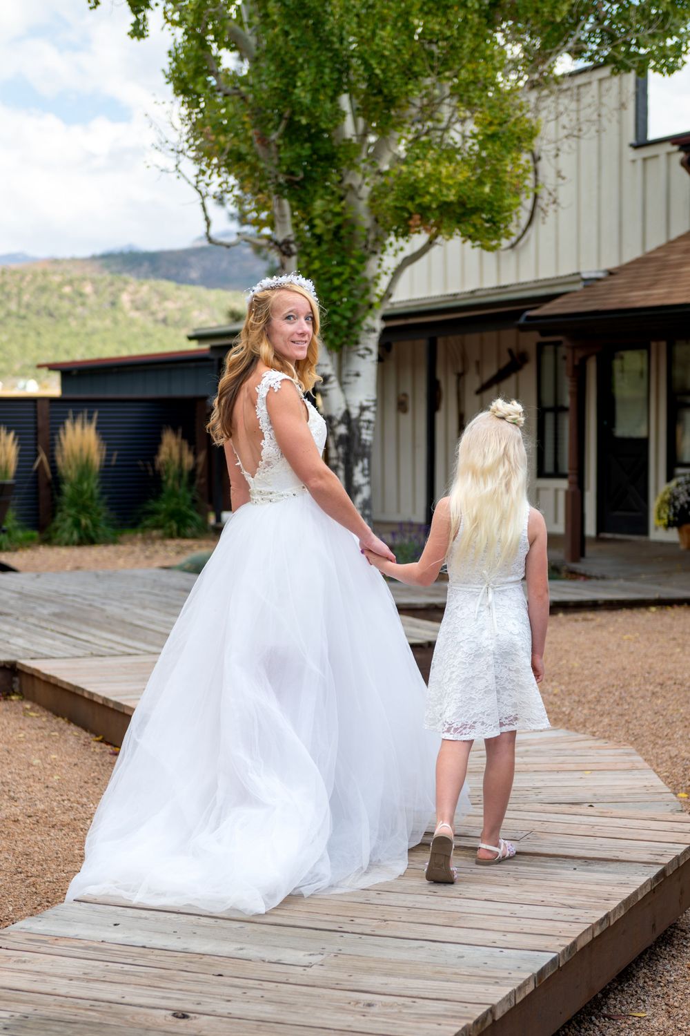 A bride is walking down a wooden pathway holding her daughters hand. She is in a white tulle gown, her daughter is in a short lace dress.