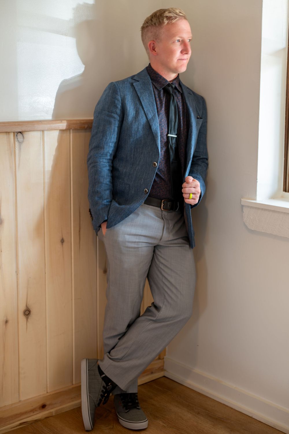 A portrait of a groom in a blue blazer and gray pants. He is standing by a window in natural light holding the lapel of his jacket.