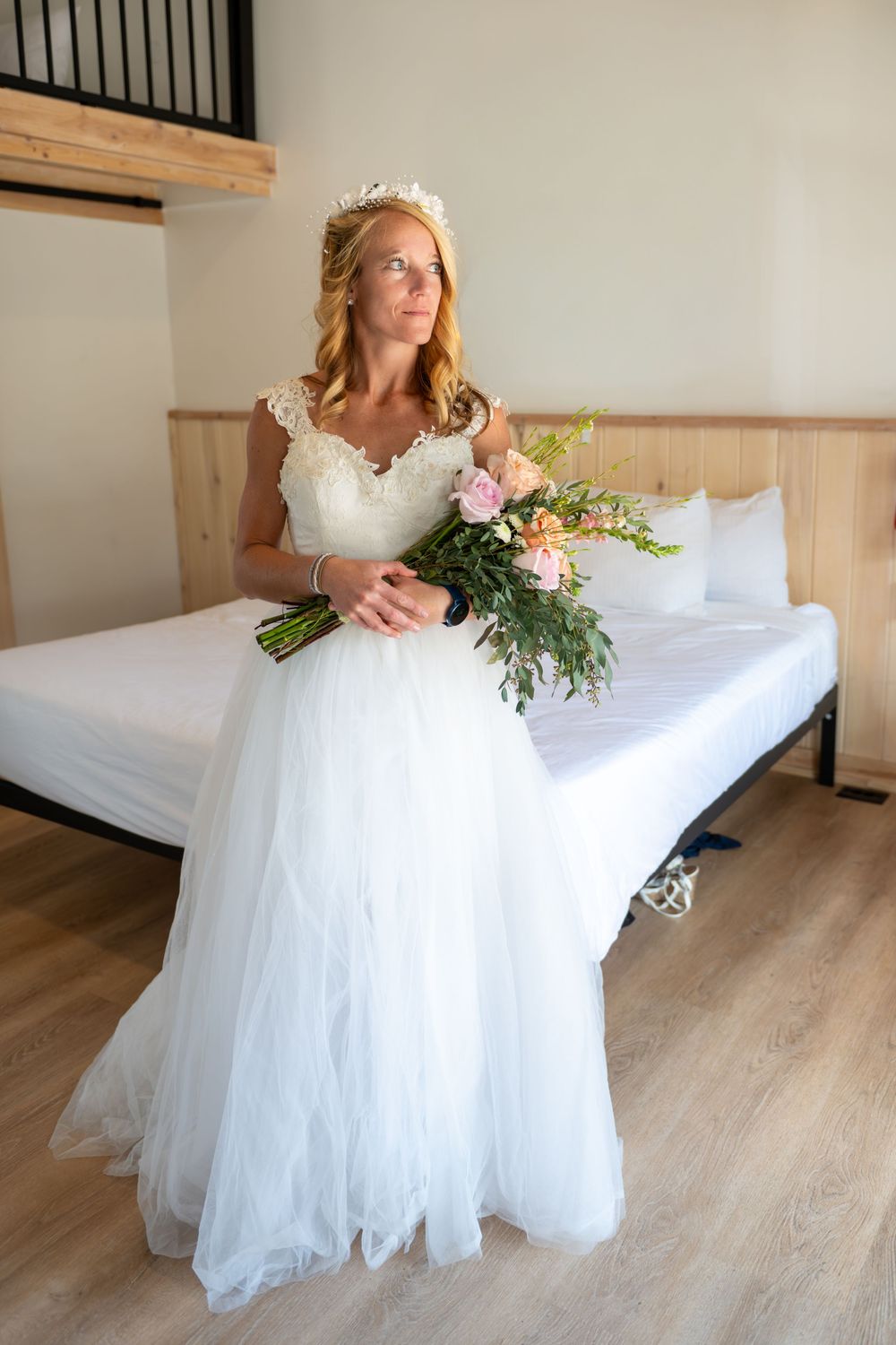 A bride in a flowing white tulle gown with lace details holds a romantic bouquet while sitting on a bed in a rustic room.