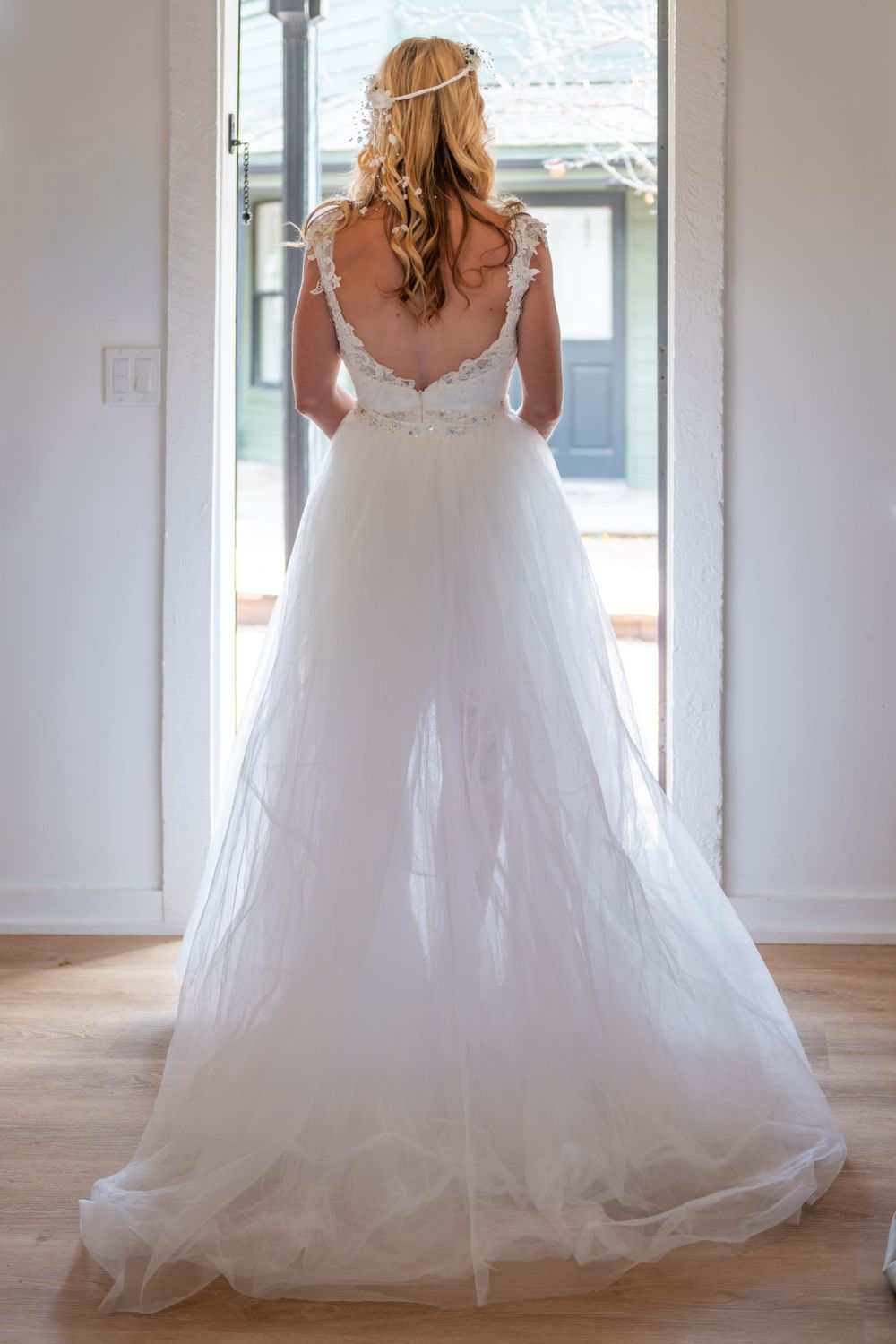 Back view of a bride standing in wedding dress with delicate lace straps and flowing tulle skirt as bride in sunlit doorway.