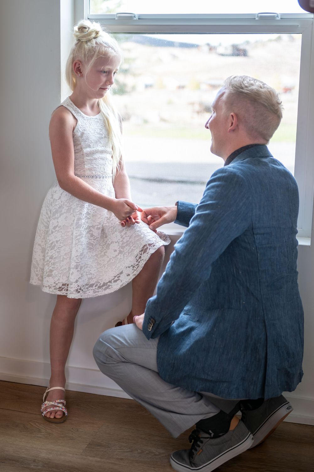 A groom kneels by his daughter in a window. She is wearing a white lace dress and strappy shoes. They are bumping fists.