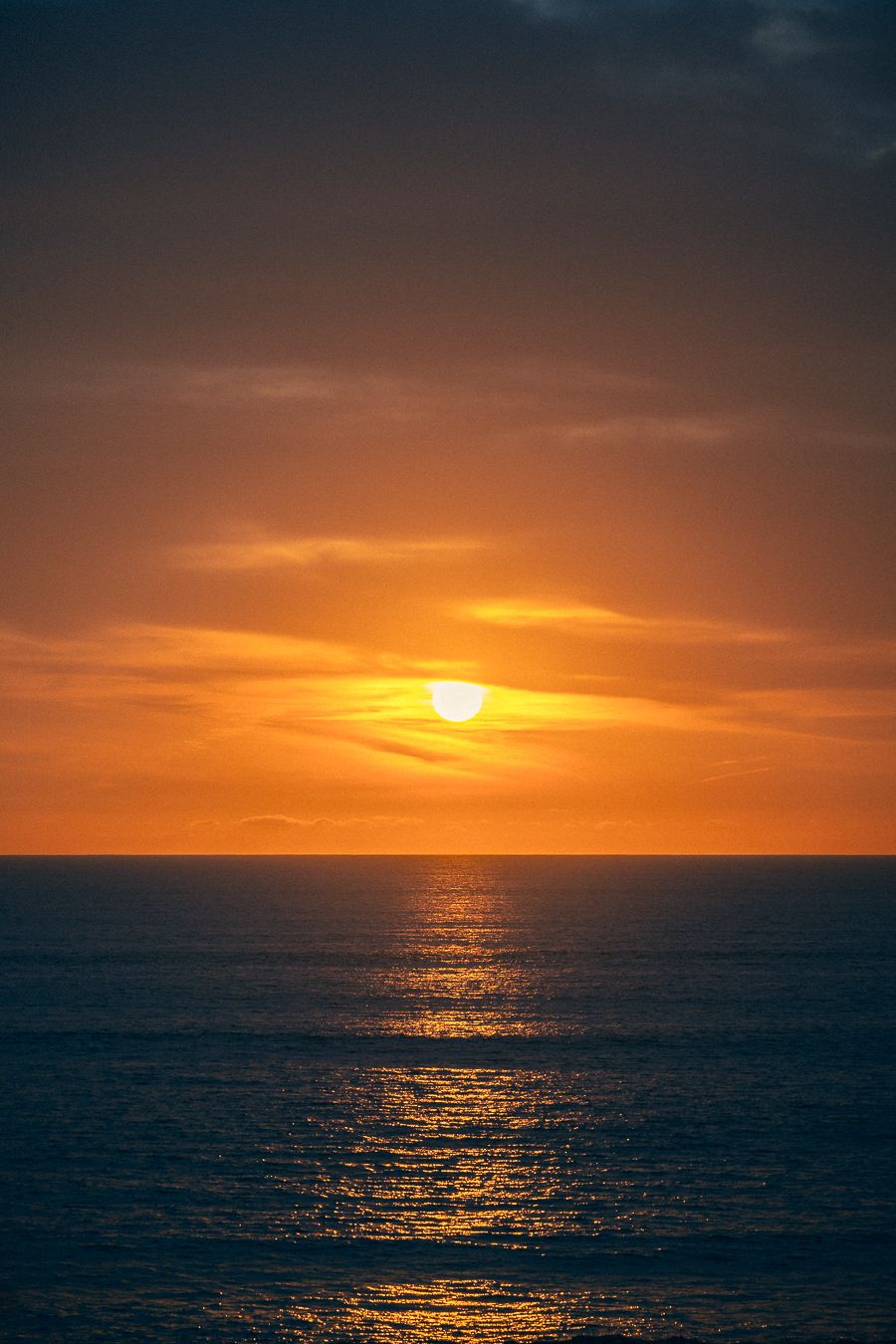 A vibrant orange sunset over calm ocean waters creates a golden reflection on the dark blue sea surface.