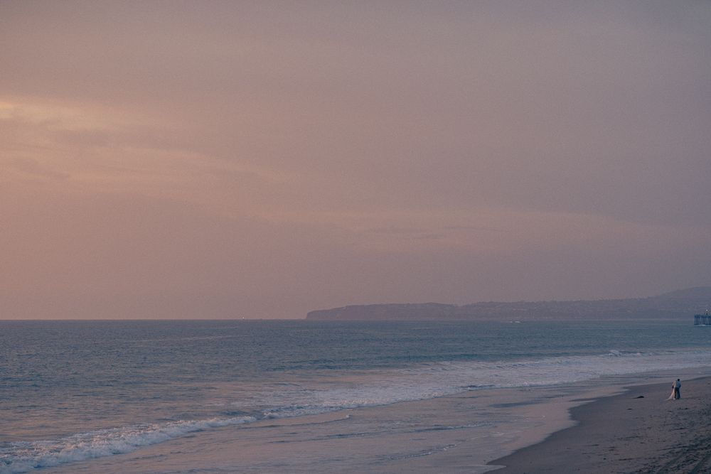 Panoramic view of a serene beach at sunset with waves rolling onto the sandy shore and distant mountains in the background.