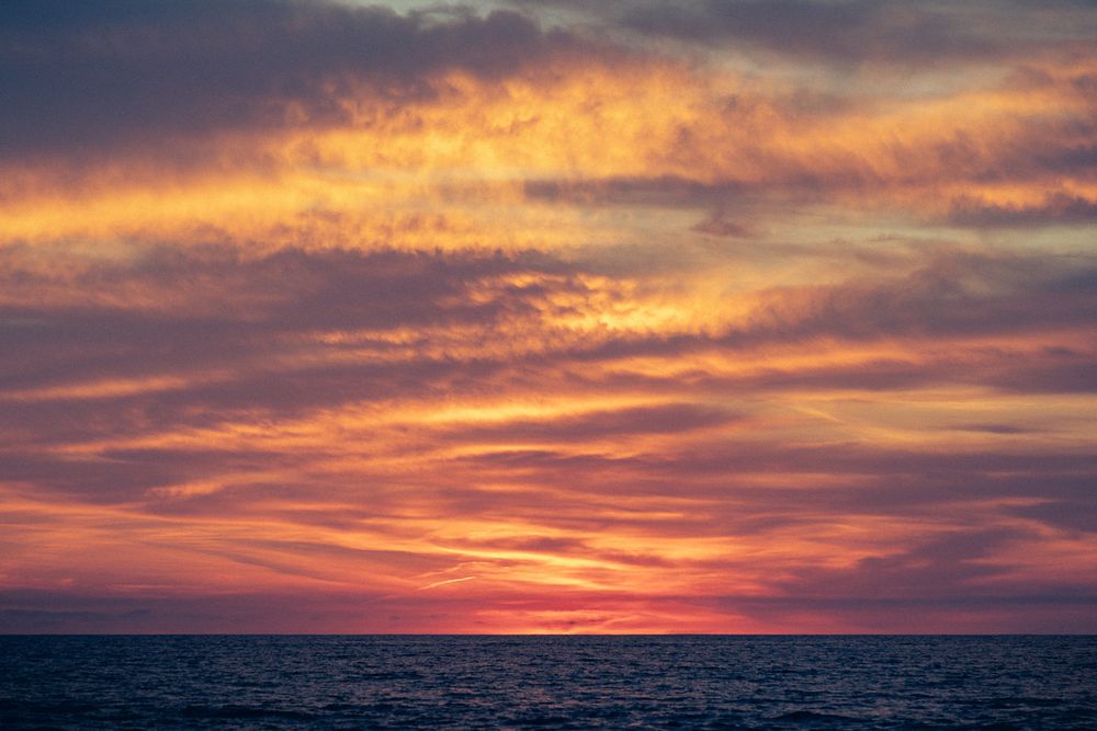 Dramatic orange and purple sunset sky over dark ocean horizon with layered clouds at dusk.