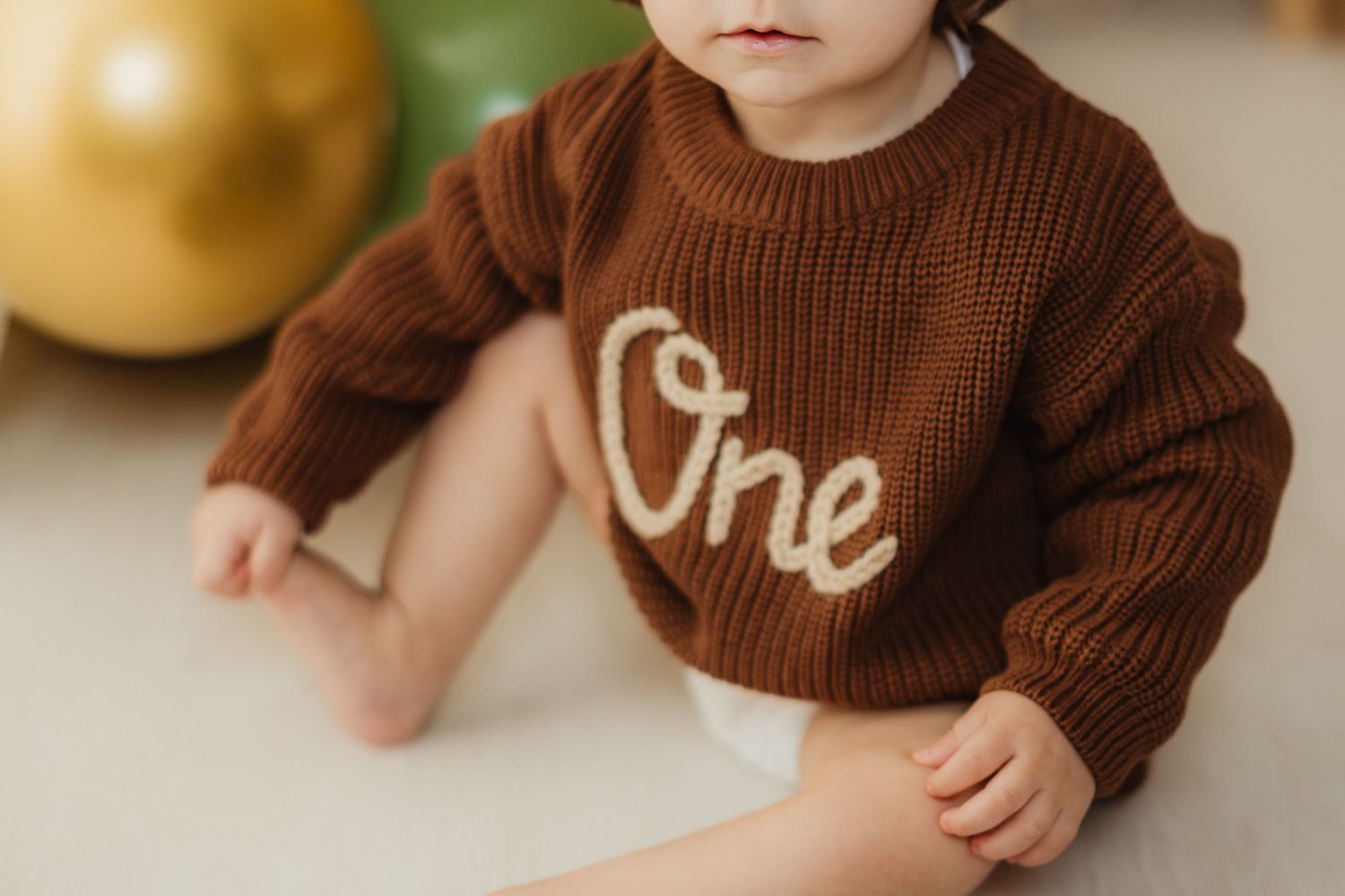 Close-up of baby wearing brown knit sweater with 'One' text near balloons.
