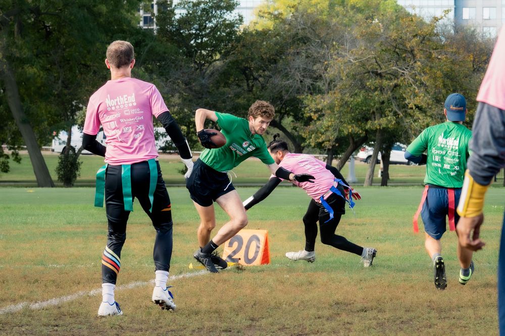 A flag football player in a green jersey with the football dodges an opponent in pink jerseys from pulling their flag in a CMSA game