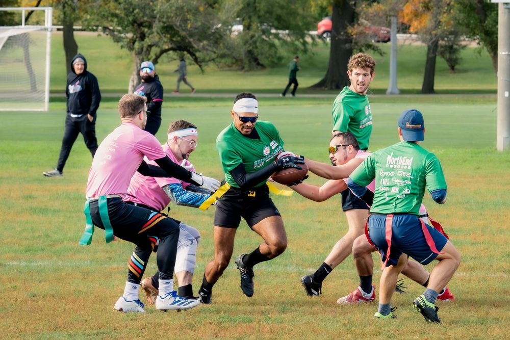 A flag football player in a green jersey with the football runs through opponents in pink jerseys in a CMSA game