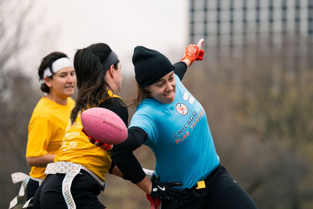 A flag football player in a blue jersey reaches forward with the football to gain extra yards while an opponent pulls their flags