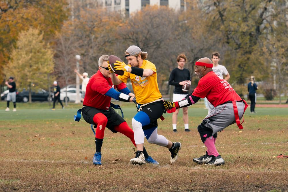 A flag football player in a yellow jersey runs downfield with the football, surrounded by opponents attempting to pull their flags