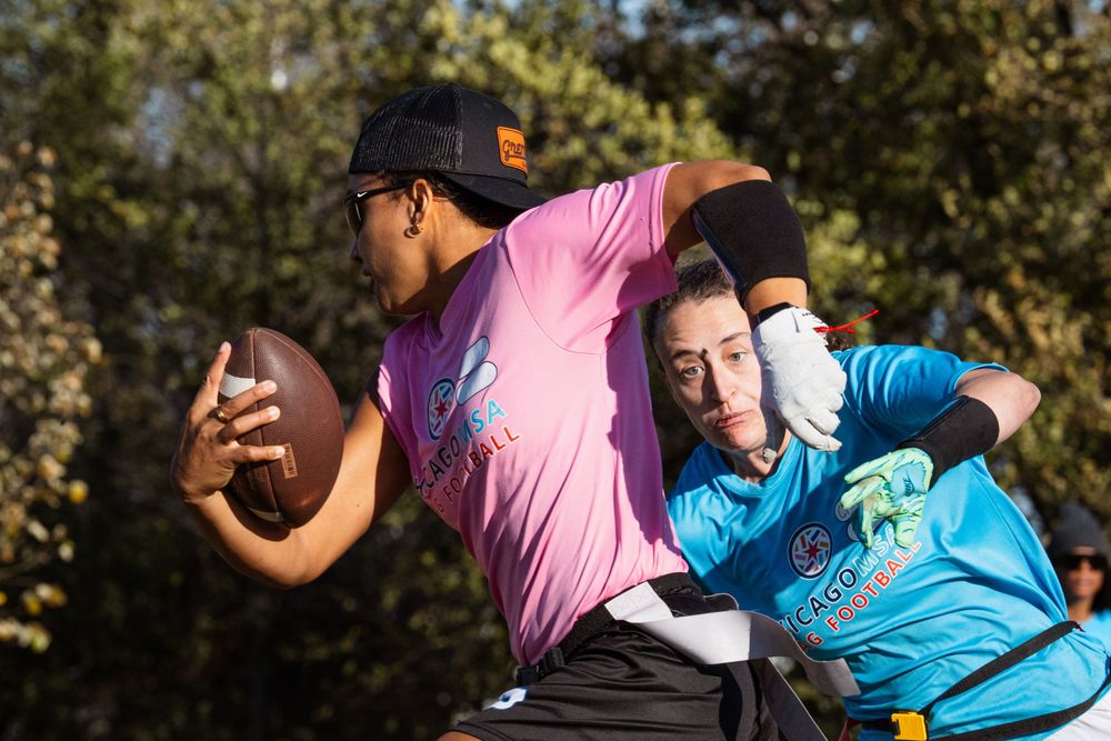 A flag football player in a pink jersey runs downfield, with an opponent in the background attempting to pull their flags to stop the play