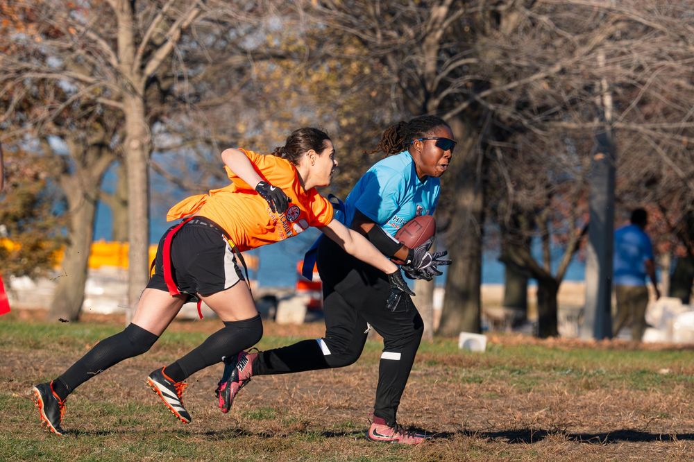 A flag football player in a blue jersey catches the football next to an opponent in an orange jersey attempting to block the pass