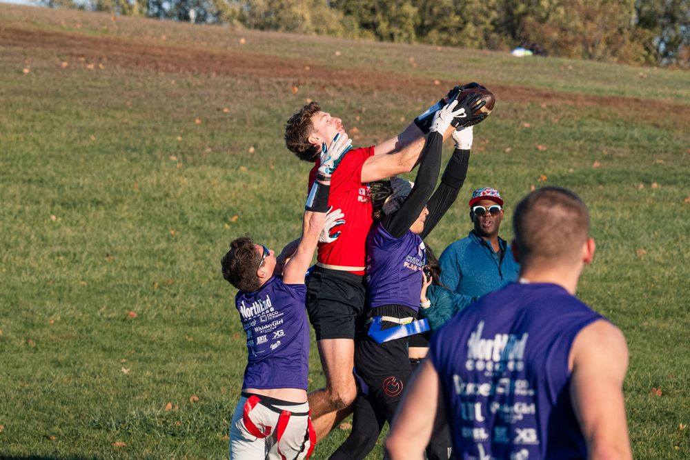 A flag football player in a red jersey catches the football in the end zone over two opponents in purple jerseys who are attempting to block