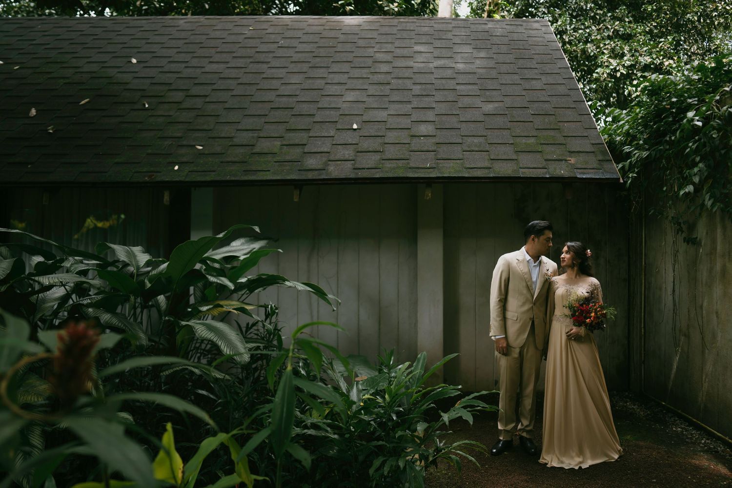 A bride and groom share a romantic moment surrounded by lush tropical greenery beside a rustic wooden building.