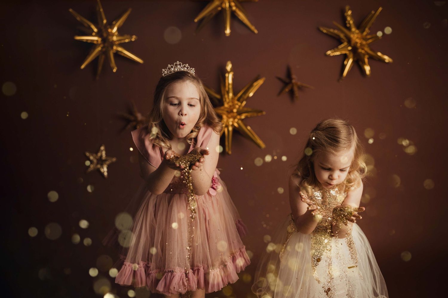 Two young children in princess dresses pose beneath golden star decorations against a dark backdrop.