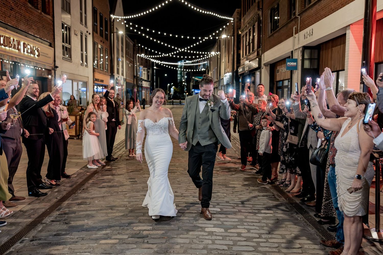 A couple walks down a cobblestone street lined with guests under twinkling lights during an evening celebration.