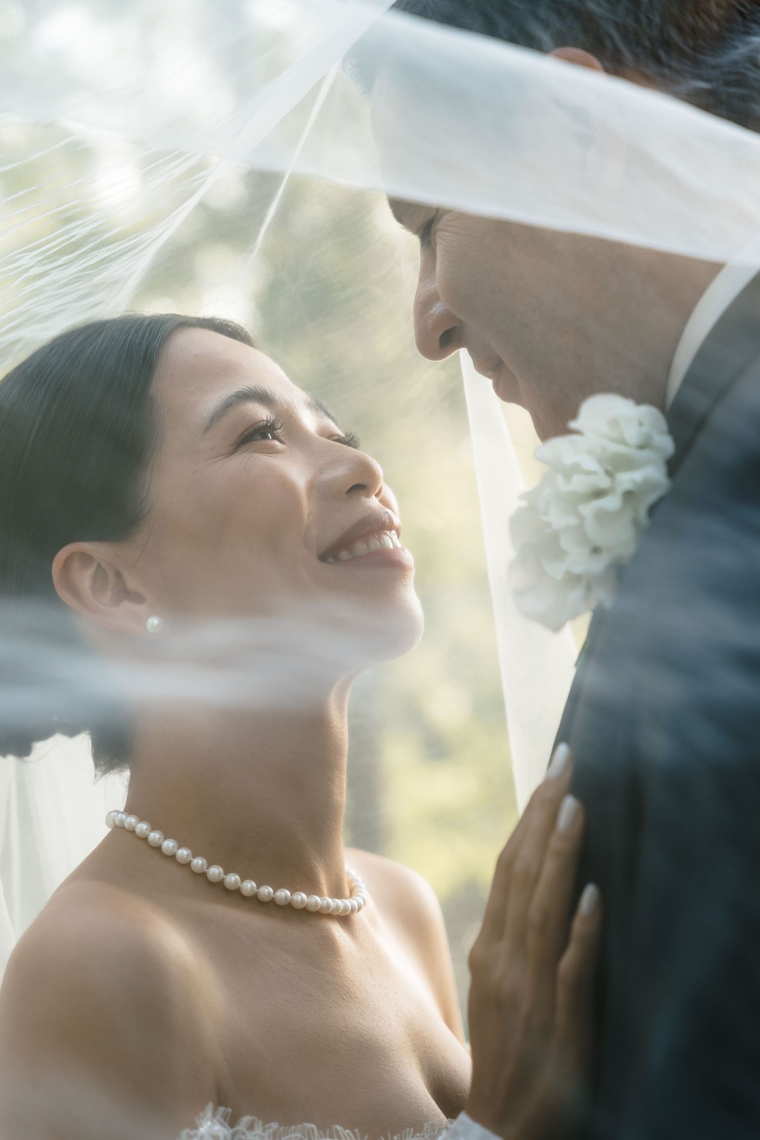 A romantic wedding couple shares an intimate moment under a soft white veil with glowing sunlight behind them.