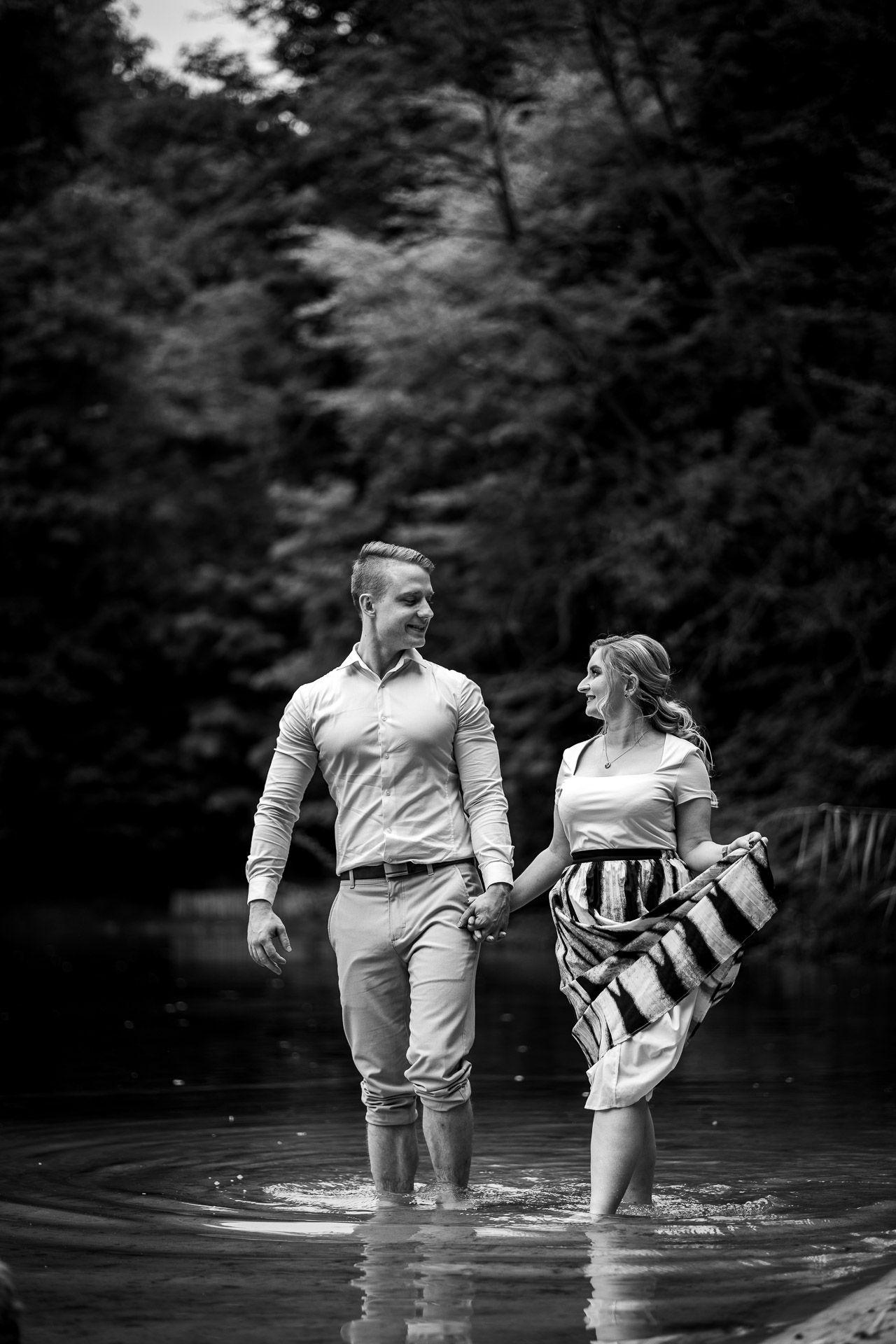 A couple wades in water together while sharing a playful moment in black and white.