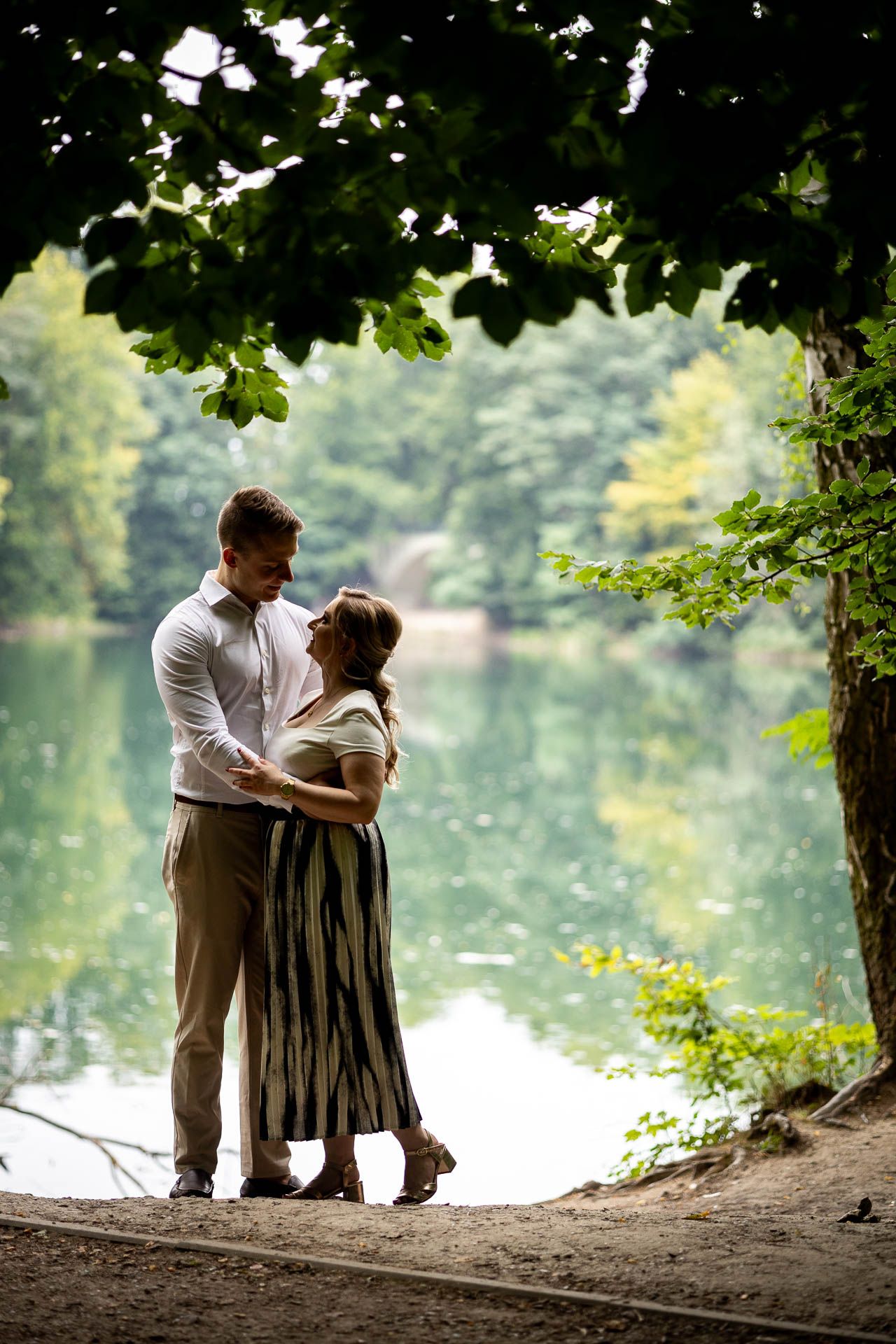 A couple stands by a tranquil lake framed by overhanging tree branches.
