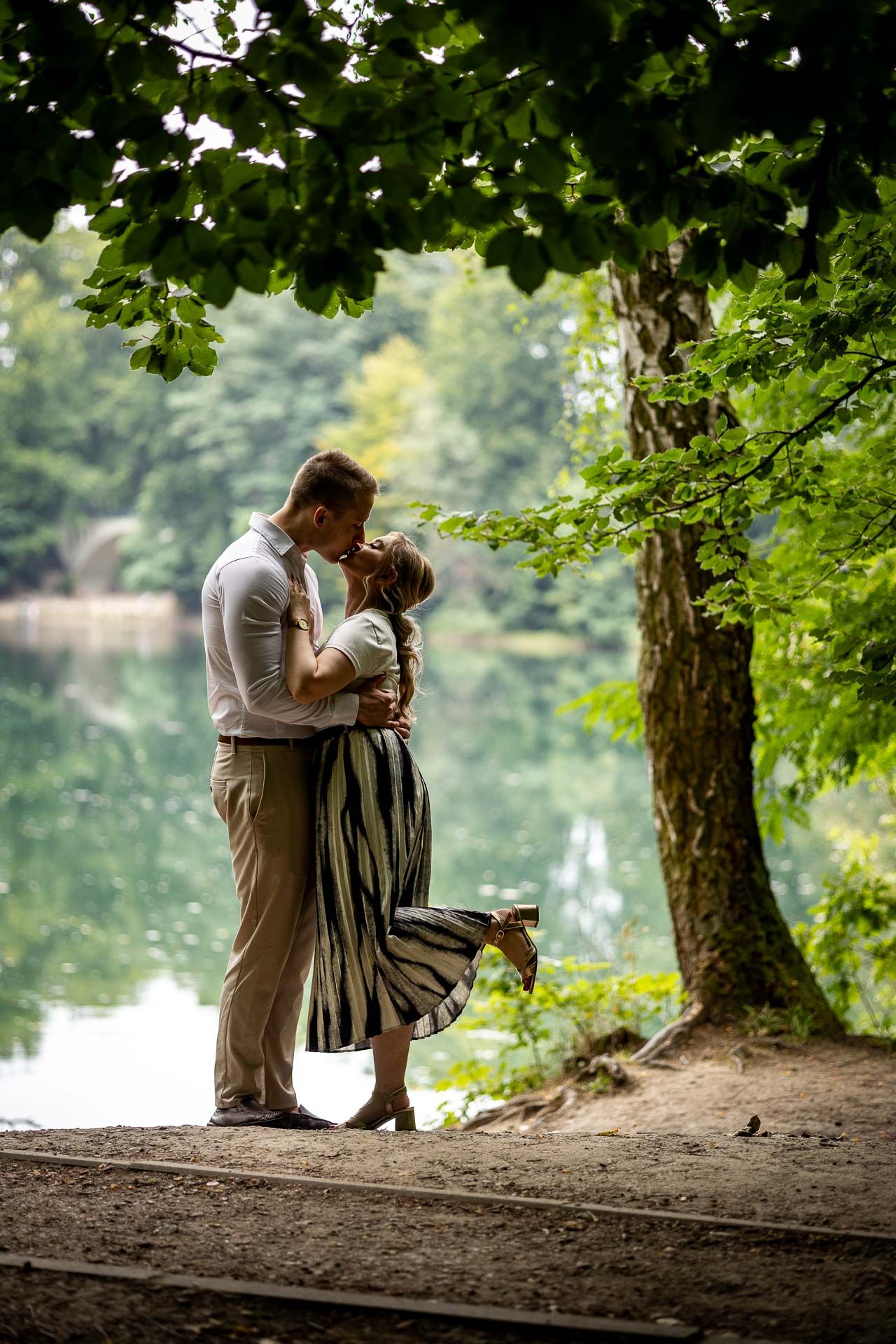 A romantic moment captured between a couple by a serene lake under green foliage.