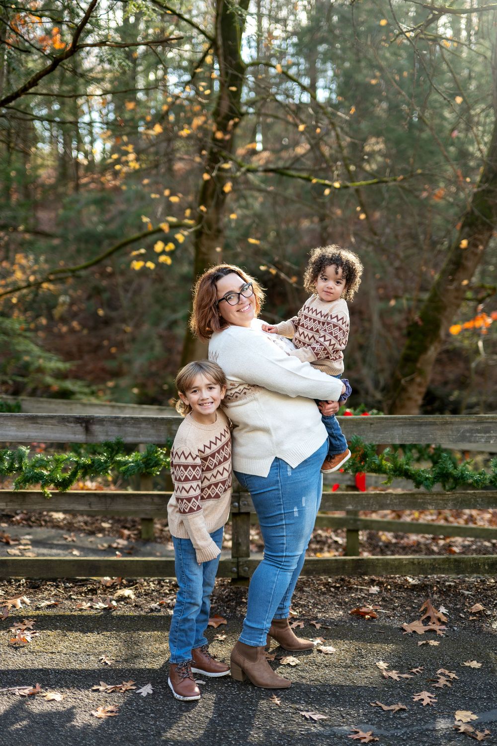 A mother holds her toddler while her young daughter stands beside her on an autumn path.