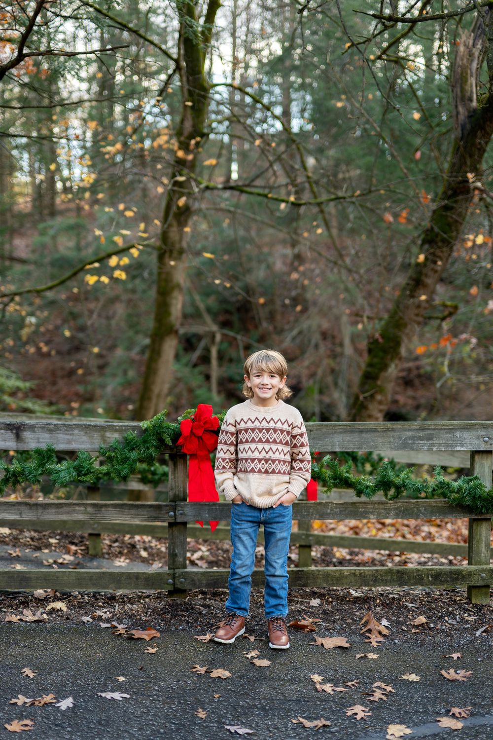 Smiling boy in fair isle sweater and jeans standing before a wooden fence decorated with Christmas wreaths in autumn forest.