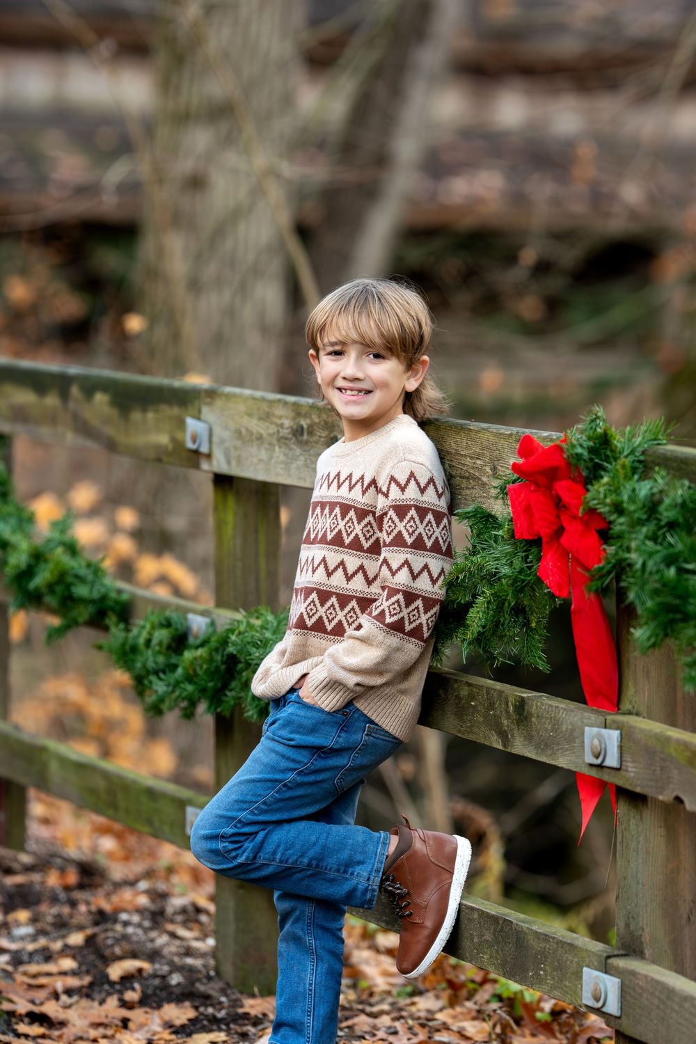 Happy boy in fair isle sweater leaning against a wooden fence adorned with Christmas garland and red bow in fall setting.