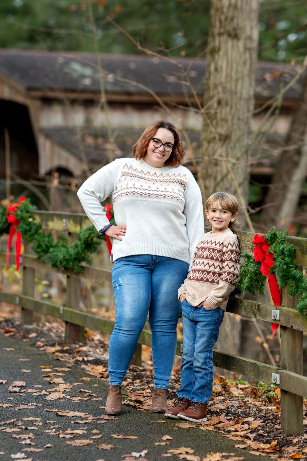 Mother and son in matching fair isle sweaters posing by a Christmas wreath-decorated wooden fence in an autumn park.