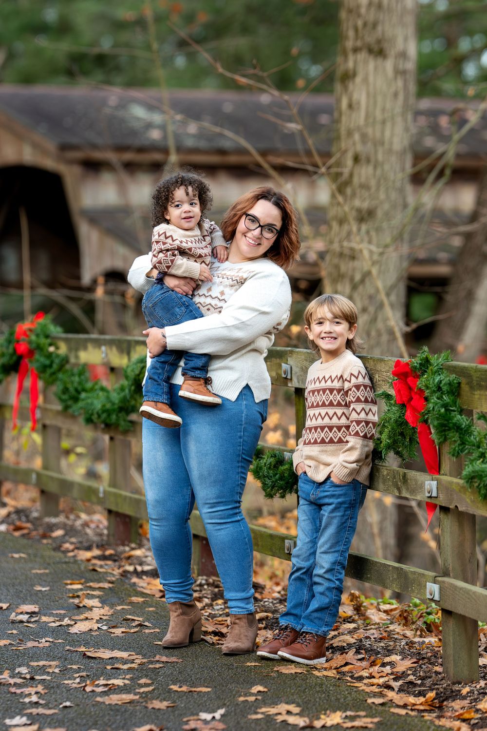 Smiling mother holding toddler while standing with older son by a Christmas-decorated fence at a covered bridge in fall.