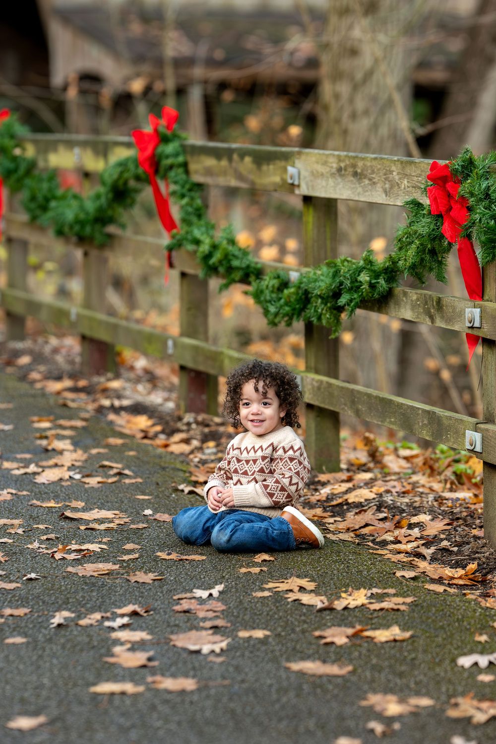 Curly-haired toddler in fair isle sweater sitting on ground near a wooden fence decorated with Christmas wreaths and red bows.