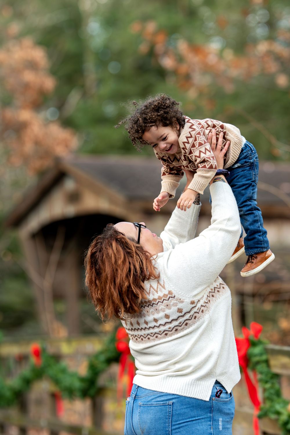Joyful mother lifting laughing curly-haired toddler in fair isle sweater outdoors near a Christmas-decorated covered bridge.