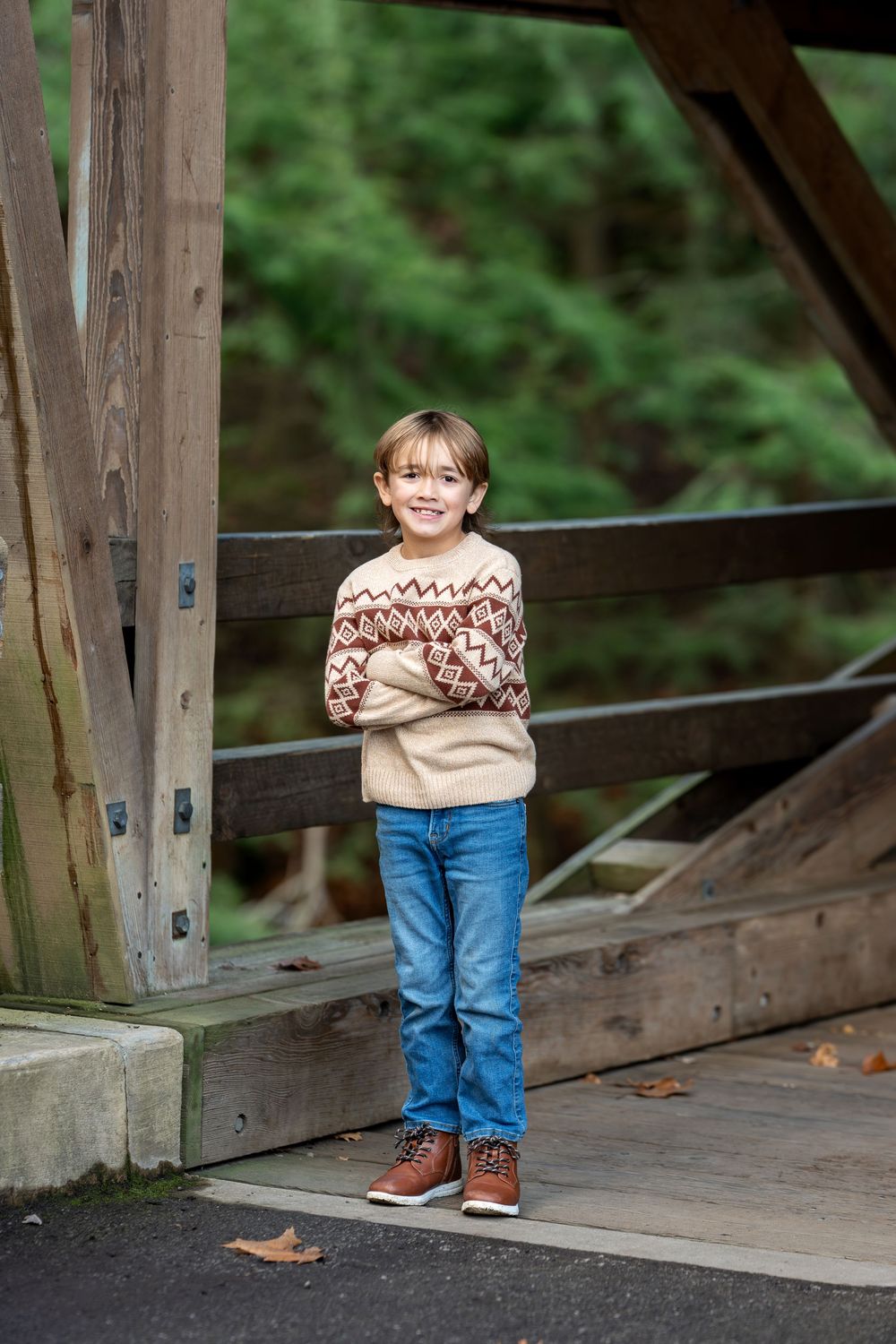 Smiling boy with glasses in fair isle sweater standing with arms crossed on a wooden covered bridge in autumn.