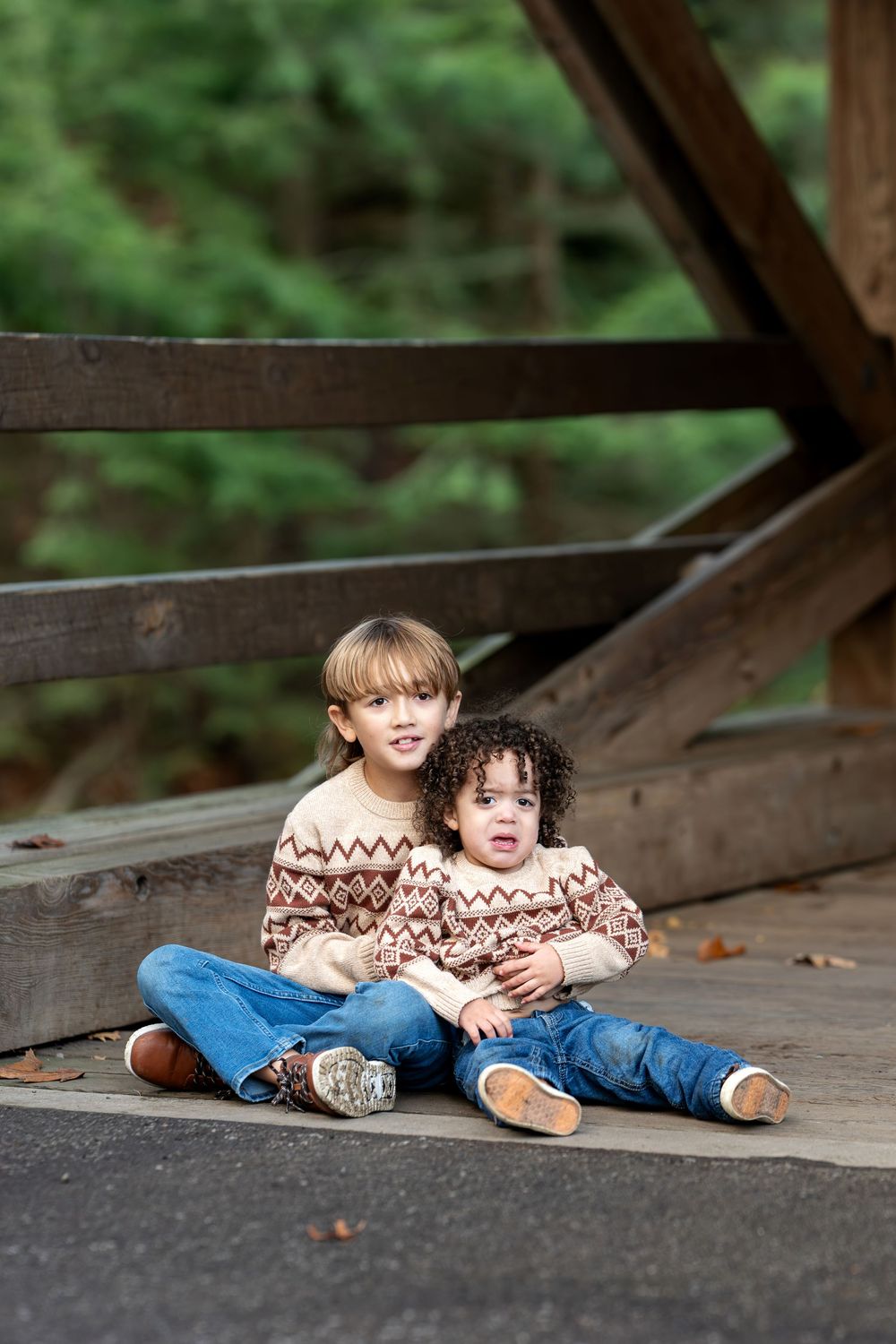 Two brothers in matching fair isle sweaters sitting together on a wooden covered bridge surrounded by green forest.